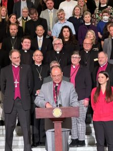 A group of people stands on steps with a man in clerical attire speaking at a wooden podium.