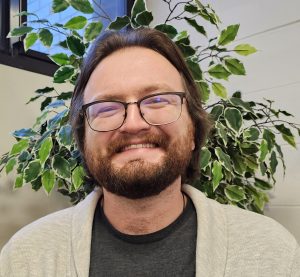 Person with glasses smiling in front of a plant indoors.