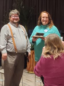 Two people smiling, one holding an award, with a person in the foreground taking a photo.