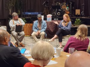 Three people are seated in a panel discussion while audience members observe.