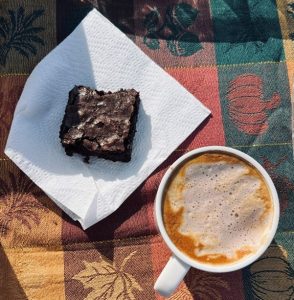 Brownie on a napkin next to a cup of frothy coffee on a patchwork autumn-themed tablecloth.