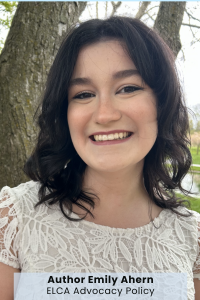 A person with dark hair and a white lace top smiling outdoors near trees.