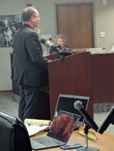 Man in a suit speaks at a podium with microphones, with a person seated at a desk behind him.