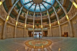 Interior of a grand rotunda with a glass-domed ceiling, marble columns, and a central floor emblem.