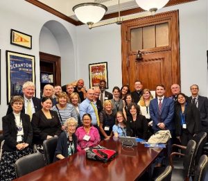 A large group of people gathered in a conference room with poster art and a wooden table.