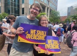 Two people holding "We Shall Overcome" signs at a rally with a crowd in the background.