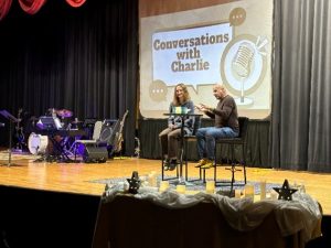 Two people seated at a stage event called "Conversations with Charlie," with musical instruments nearby.