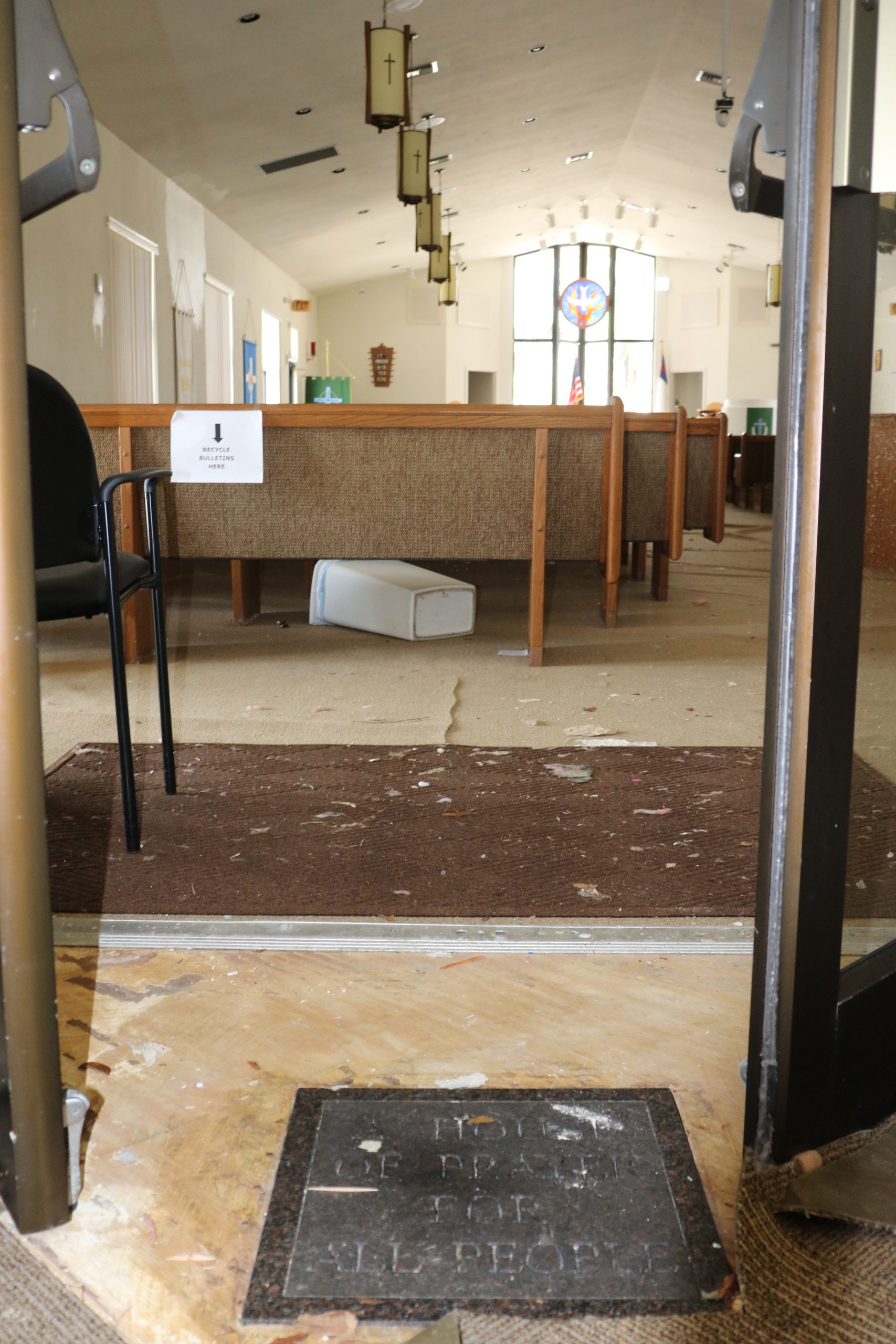 The entrance to a church sanctuary. There is debris across the floor and back pews. At the door threshold, a stone plaque in the ground reads "House of Prayer for All People."