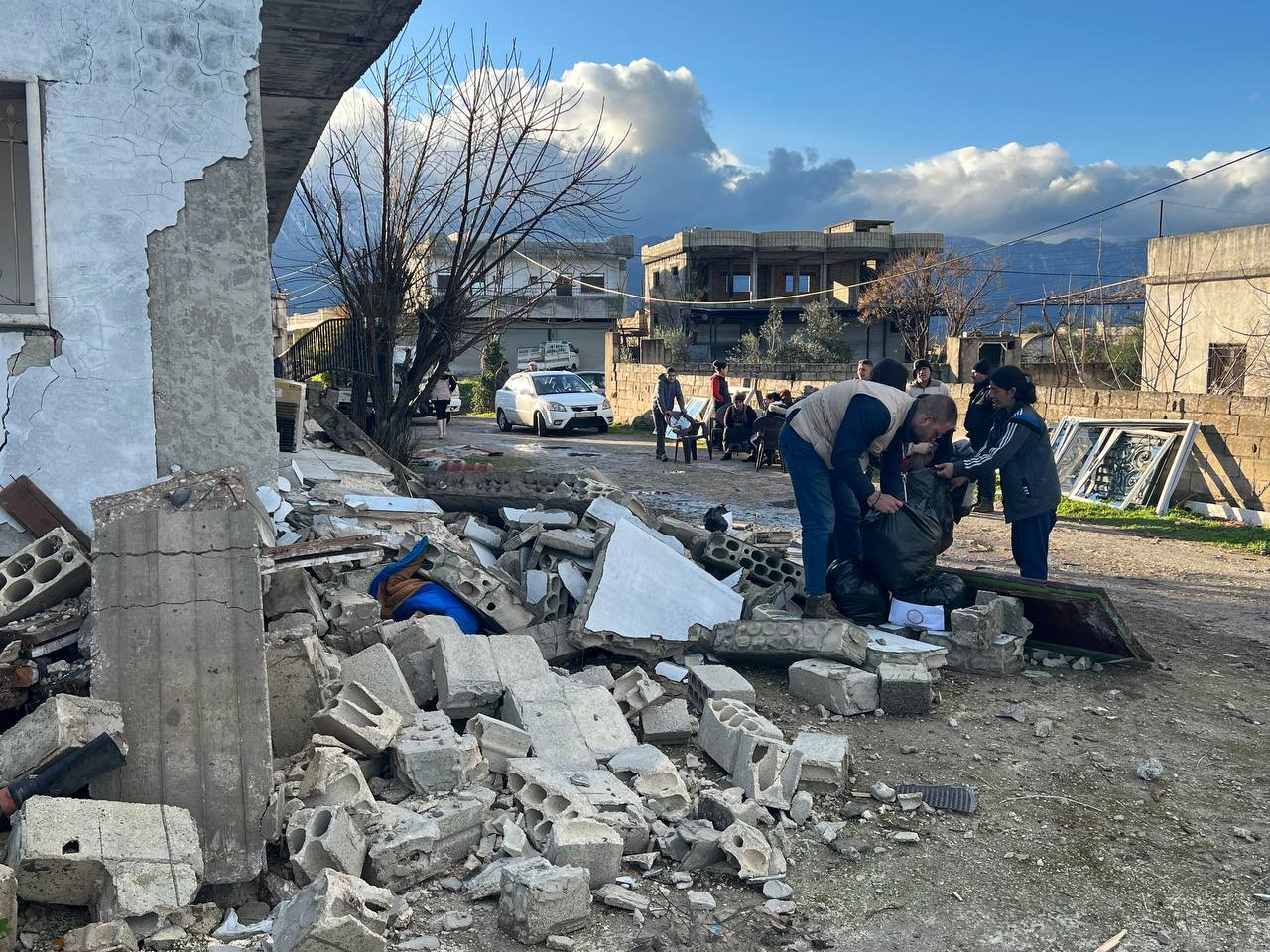 On the left, a crumbled building. On the right side of the building are two people picking up the rubble.