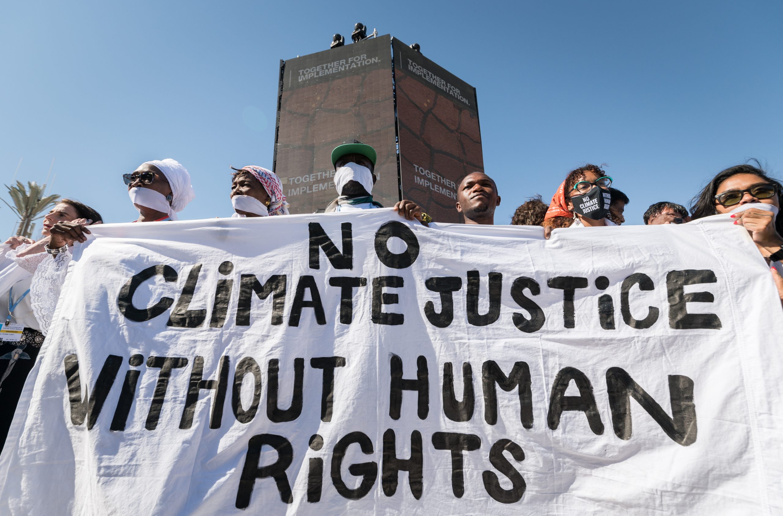 A group holding a banner reading 'No Climate Justice without Human Rights'. The group is pictured in front of a large pillar with the COP27 slogan 'Together for Implementation' on display.