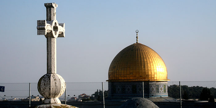 Image of the Dome of the Rock in Jerusalem, foregrounded by a cross. Both the cross and the Dome of the Rock are separated by a chain-link fence.
