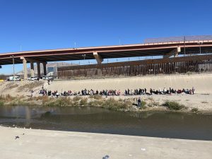 A river cuts the foreground, while a line of people gathers in front of an opening in the bridge.